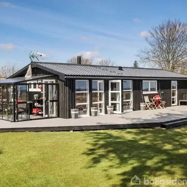 Sort træhus med store vinduer står på en træterrasse. Vedhæftet er en glaseret udestue. Græsklædt have omgiver huset, og himlen er blå med få skyer.