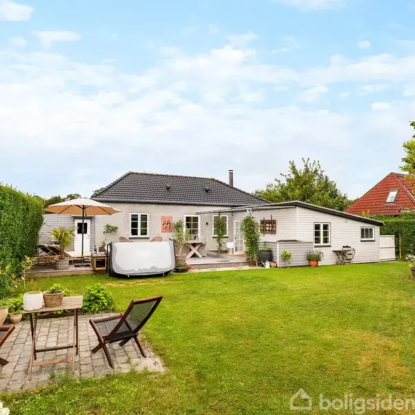 Et hus med gråt tag og hvidmalet facade ligger i en velholdt have. En terrasse har havemøbler og parasol, mens græsplænen rummer haveborde og stole omgivet af hække og planter.