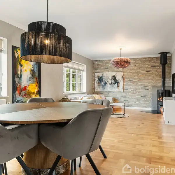 Spisebord med grå stole står i en stue. En stor, sort lampe hænger over bordet. Væggene er dekoreret med malerier, og der er en brændeovn i baggrunden.
