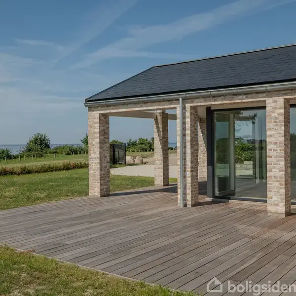 En moderne terrasse med trædæk strækker sig fra et hus med murstensmure og store vinduer. Haven omkring er velplejet med udsigt over græs og vand under en klar blå himmel.