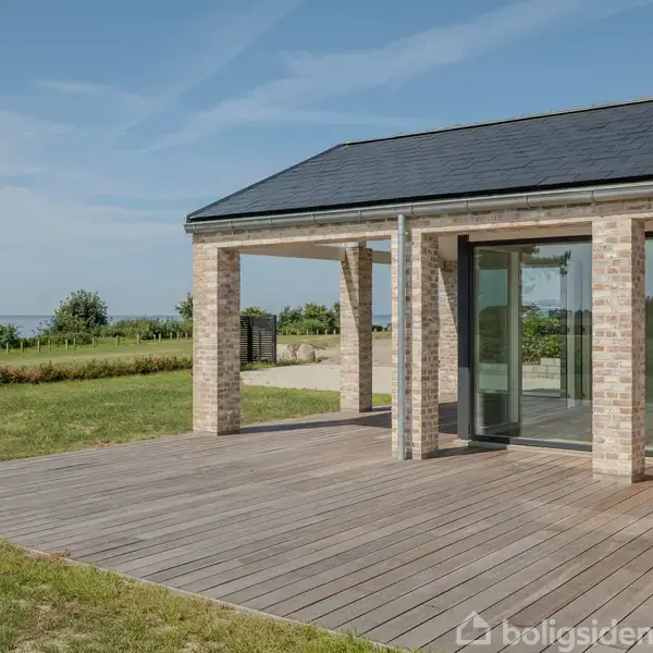 Hus med store vinduer ligger på en træterrasse, omkranset af en grøn græsplæne. Baggrunden viser havudsigt under en klar blå himmel.