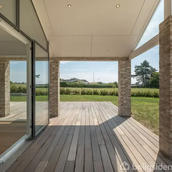 En overdækket terrasse med trægulv strækker sig ud fra et moderne hus med store vinduer. I baggrunden ses en grøn græsplæne og træer under en klar himmel.