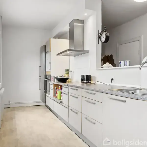 Køkken med hvide skabe og grå bordplade; vask og kogeplader findes under emhætte. I baggrunden ses et vindue og en radiator. Køkkenet har lyst trægulv og vægge.