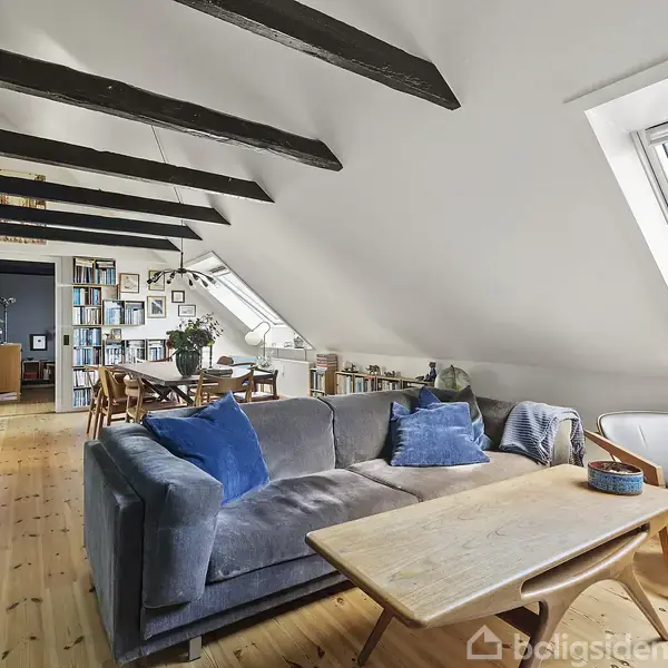 En stue med en grå sofa med blå puder står ved et træbord. Skråt loft med synlige bjælker. Store skråvinduer slipper lys ind, og bøger fylder reolerne bagved.
