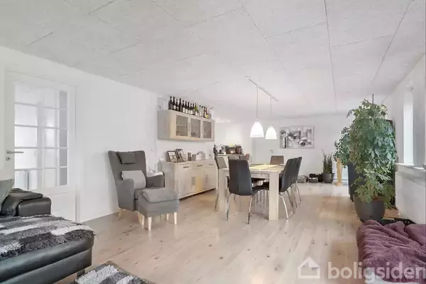 Stue med spisebord og stole centreret i midten, omgivet af lænestol og sofa. På væggen er der en skænk med bøger og flasker ovenpå. Store vinduer giver naturligt lys.