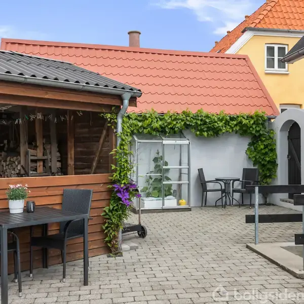 En gårdhave med borde og stole står på en brolagt terrasse omgivet af bygninger med farverige tage. Et træskur er fyldt med brænde, og planter klatrer op ad væggen.