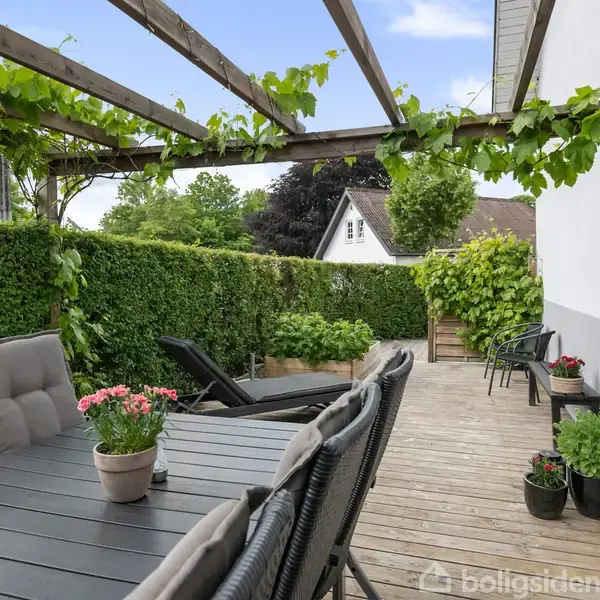 Havebord med grå stole står på en træterrasse. Over bordet hænger en pergola med grønt løv. I baggrunden ses hække og planter i en have.