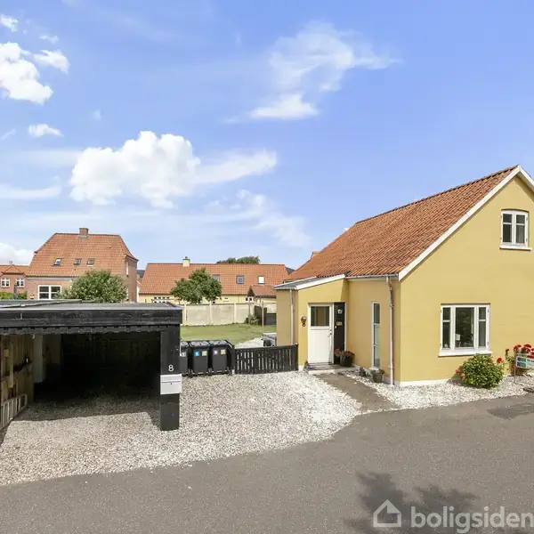 Gul hus med rødt tag står ved siden af en sort carport på grusbelagt grund. Blå himmel med skyer og flere lignende huse i baggrunden. Flagstang med dansk flag.