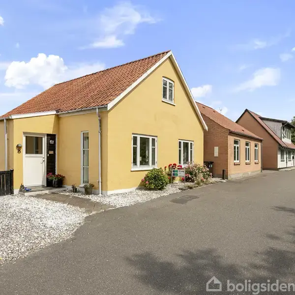 Et gult hus med rødt tegltag står ved en asfalteret vej, omgivet af en hvid grusindkørsel og grønne buske under en blå himmel med skyer.