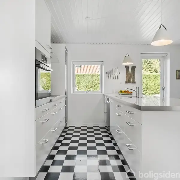 Et lyst og moderne køkken med sort-hvid ternet gulv, hvide skabe og skuffer. Et vindue og dør giver udsigt til en grøn hæk. To pendellamper hænger fra loftet.