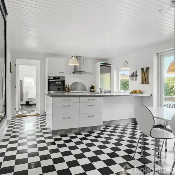 Et moderne køkken med et sort-hvidt ternet gulv. Et hvidt spisebord med stole står ved et vindue. Over køkkenøen hænger to lamper, og udenfor ses en have.
