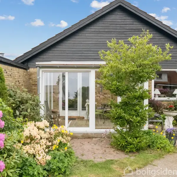 Et enfamiliehus med et glasparti og terrasse, omgivet af farverige blomster og grønne buske i en velplejet have under en blå himmel med få skyer.