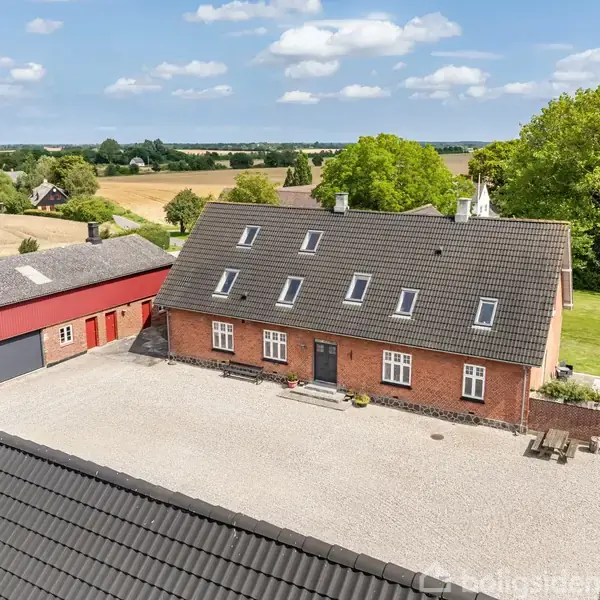 Et stort, rødt murstenshus står stille på en gårdsplads omgivet af grus og grønne marker i baggrunden under en blå himmel med spredte skyer.