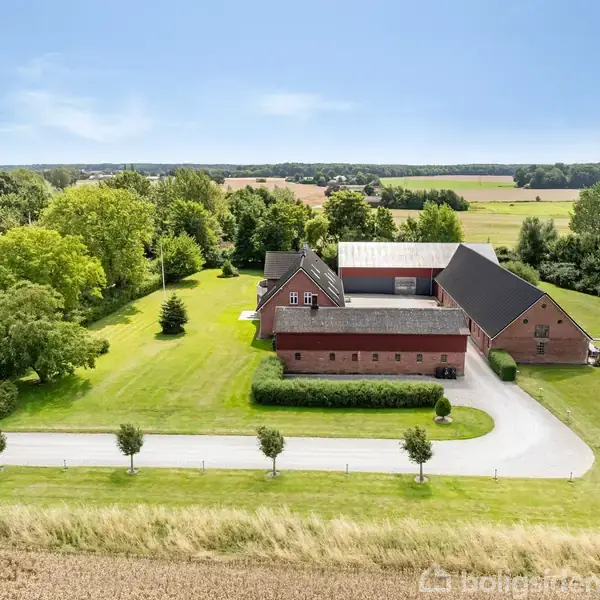 En stor landbrugsejendom i røde mursten ligger stille på en velplejet grøn grund omgivet af træer og marker under en klar blå himmel.