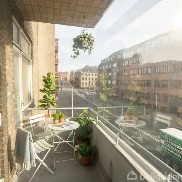 En altan med et lille rundt bord og to stole, planter omkring, hænger over en travl byvej. Omringet af moderne murstensbygninger.