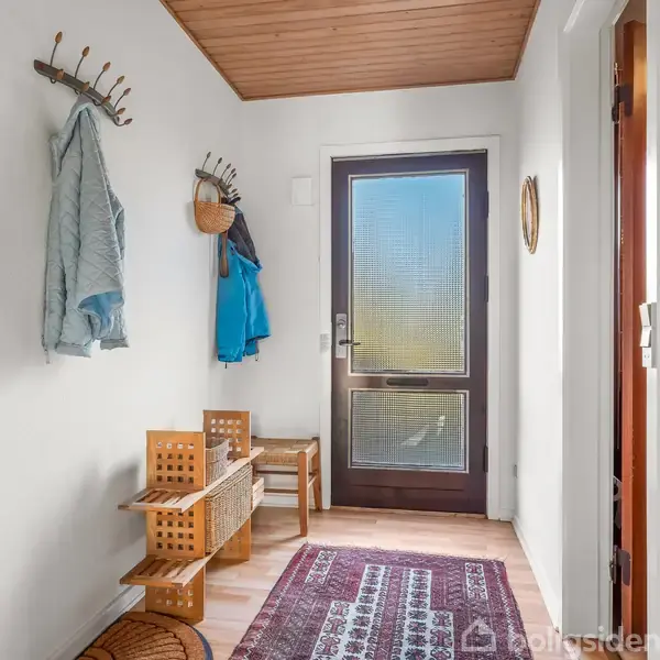 Jakker hænger på en knagerække, mens en skohylde står ved siden af en indgangsdør i et rummeligt, lyst entréområde med trægulv og væg-til-væg tæppe.