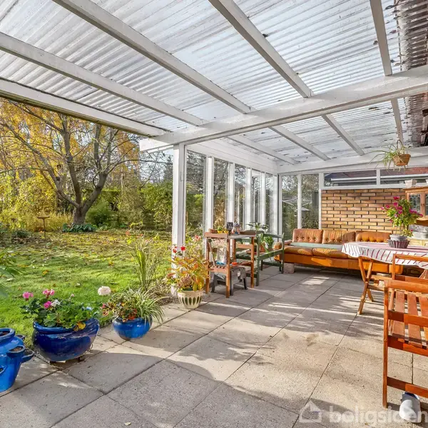 Terrasse med klare tagpaneler og brune havemøbler. Blå blomsterpotter og grønne planter omkranser området. I baggrunden ses en frodig have med træer og buske i efterårsfarver.
