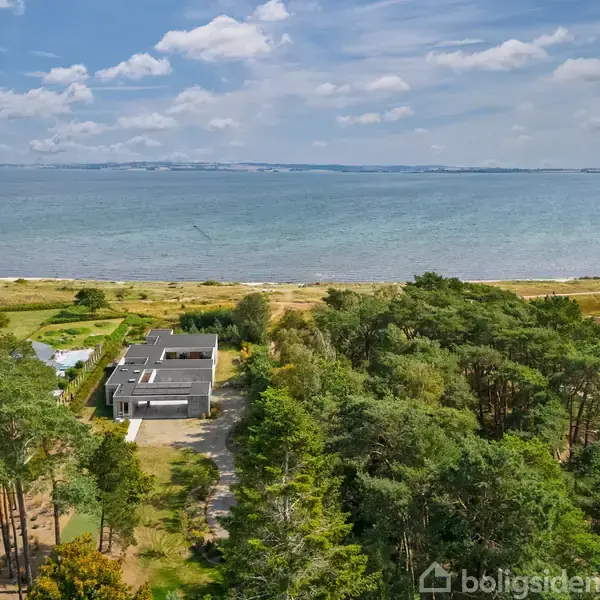 Et moderne hus ligger stille omgivet af skov tæt på en strand med udsigt over havet, under en himmel med spredte skyer.