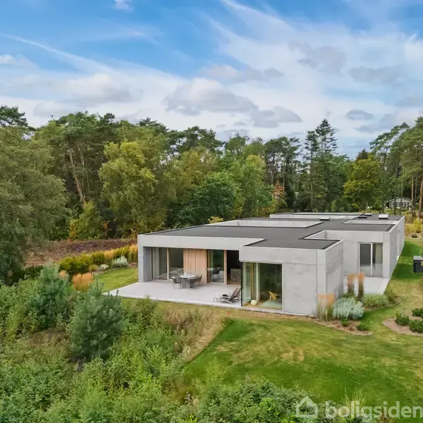Et moderne, minimalistisk hus ligger omgivet af frodig natur og træer. Huset har store vinduer og en terrasse, der åbner op til en velplejet græsplæne.