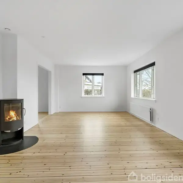 Brændeovn fyret op, placeret i et rummeligt, lyst værelse med trægulv. Store vinduer på højre væg slipper lys ind, og der er en radiator under det ene vindue.