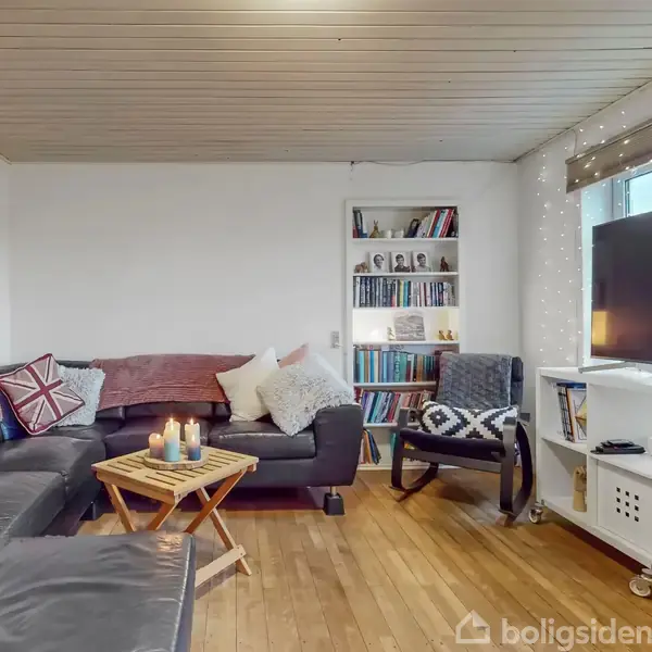 En stue med sort hjørnesofa dækket af puder, står ved siden af et sofabord med stearinlys. En reol og tv er placeret i rummet med trægulv og lyse vægge.