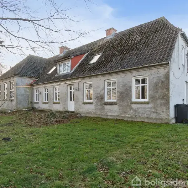 En stor, ældre bygning med hvidkalket facade og sort tegltag står på en græsplæne omgivet af træer og åben himmel. Der er ingen tekst på billedet.