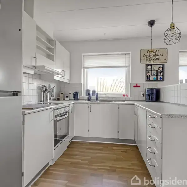 Køkken med hvidt køkkenskab og stålgreb, ovn og opvaskemaskine. To vinduer bagved slipper lys ind. På væggen hænger tekstskilt: "Please excuse the mess ... children are making memories.".