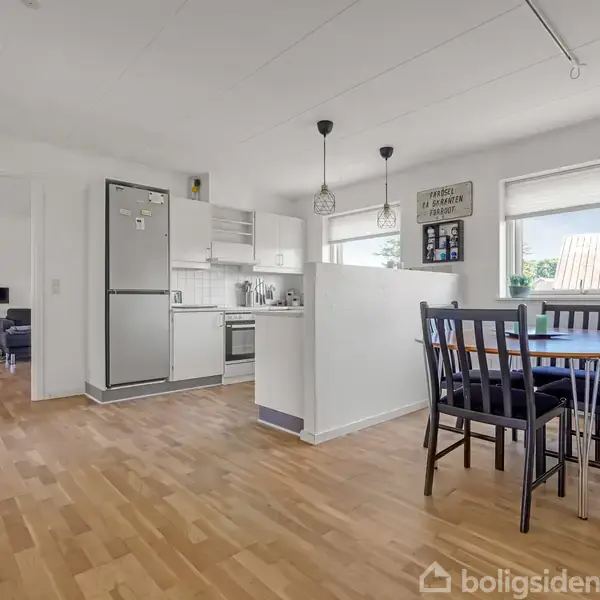 Spisebord med stole står ved siden af et åbent køkken i et lyst rum med trægulv. På væggen hænger kunst og skilte, der skaber en hjemlig atmosfære.