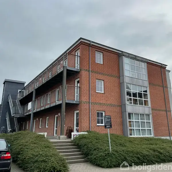 En rød murstensbygning har altaner og trapper op langs siden. En parkeret bil står foran på en stigende adgangsvej. Skyerne hænger lavt i baggrunden.