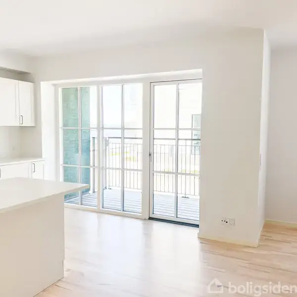 En lys og moderne køkken-alrum med hvide elementer, en køkkenø med vask. Store glasdøre leder ud til en balkon. Parketgulve og hvide vægge skaber en minimalistisk atmosfære.