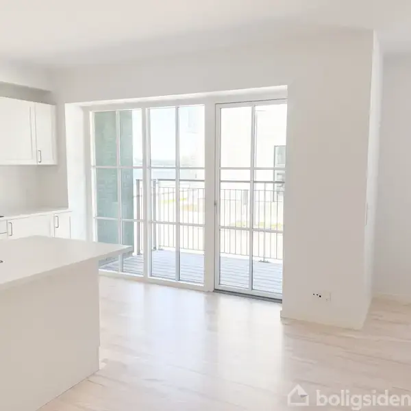 Køkken med hvidt inventar og sort kogeplade ved siden af en stor glasdør, der åbner ud til en altan. Rummet er lyst med lyse trægulve.