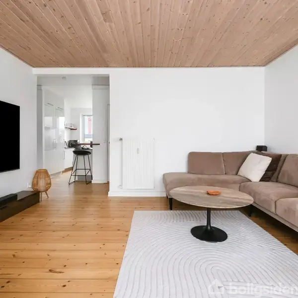 En beige hjørnesofa står i en stue med trægulv og lyst træloft. Foran sofaen er et rundt sofabord på et hvidt tæppe. Til venstre hænger et stort TV på væggen. I baggrunden ses en åbning mod et køkkenområde.