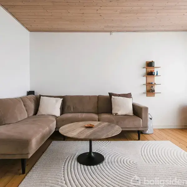 En brun hjørnesofa står mod en hvid væg i en stue. Foran er et rundt sofabord, og ved siden er en globusbar. Der hænger en lille hylde med dekorationer.