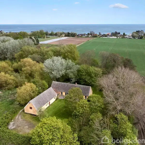 Et aflangt gult hus med stråtag ligger omgivet af tætte grønne træer i et landligt område. I baggrunden ses åbne marker og kystlinjen ved havet.
