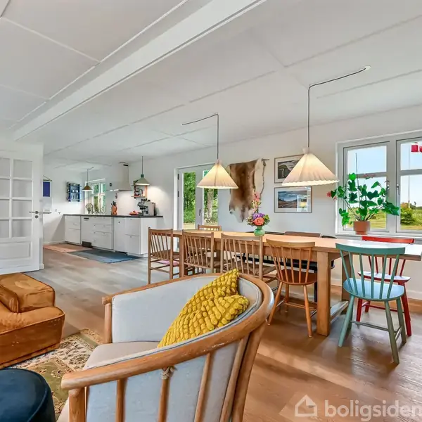Stue med sofa og stole, der står omkring et tæppe. En spisestue med et langt bord og stole ses ved vinduet. Køkken i baggrunden. Lysindfald fra store vinduer.
