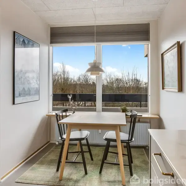 Et lille spisebord med to stole står ved et vindue. Udenfor kan man se træer i det fjerne. Rummet er lyst og enkelt indrettet.