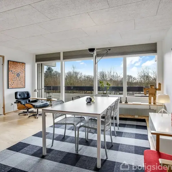 Et spisebord står i en lys stue med store vinduer, omgivet af moderne stole, en lænestol og kunst på væggene. Udenfor ses trætoppe under en blå himmel.