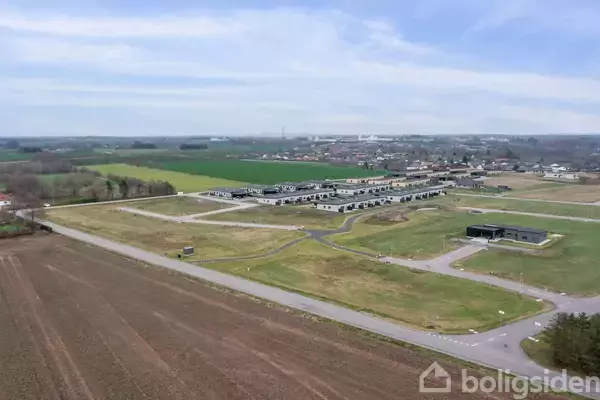 En stor mark med enkelte nybyggede huse ses fra luften. Husene er omgivet af åbne marker og veje, med en by i baggrunden og en klar, blå himmel.