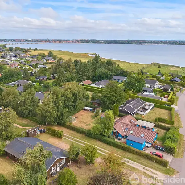 Huse ligger spredt i et grønt område ved vandet. En vej snor sig gennem kvarteret, og træer omkranser bygningerne. I baggrunden ses en bred udsigt over vandet og horisonten.
