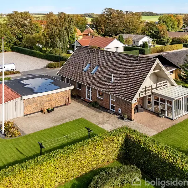 Hus står stille med stor have og græsplæne omgivet af hække. Der er en udestue og en garage ved siden af. Omkringliggende huse kan ses i baggrunden.
