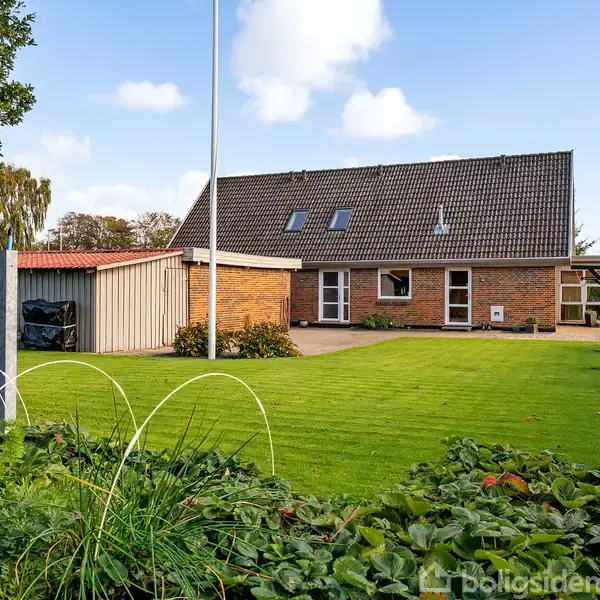 Hus med røde mursten og gråt tag står i en velplejet have med græsplæne og små køkkenhavebede. Ved siden af er en grå carport; himlen er let skyet.