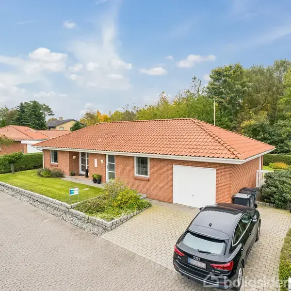 En rød murstensvilla med rødt tegltag står stille på en brostensbelagt vej. En sort bil er parkeret i indkørslen. Omgivet af grønne buske og en velplejet græsplæne.