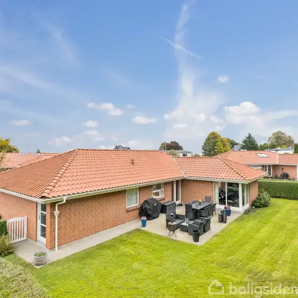 Et rødt murstenshus med rødt tegltag står stille i en velplejet have med hæk. Havemøbler og en grill står på en terrasse i den solbeskinnede baggård.