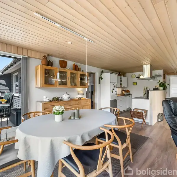 Spisebord med seks stole er centralt placeret i en lys spisestue. Rummet har åbent til et køkken og udsyn til en udendørs terrasse gennem store glasdøre.