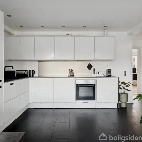 Et moderne køkken er indrettet med hvide skabe, en indbygget ovn og mørkt gulv. Til højre står en grøn plante ved siden af en åbning til en anden stue.