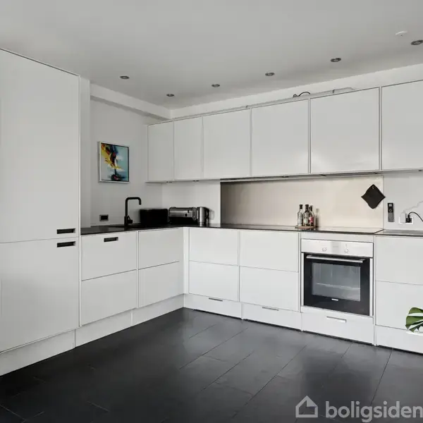 Hvide køkkenskabe flankerer en vinkelformet, moderne køkkenvæg med indbygget komfur, sort vask og kaffemaskine. Et lille maleri hænger ved vasken. Plante på gulvet til højre.