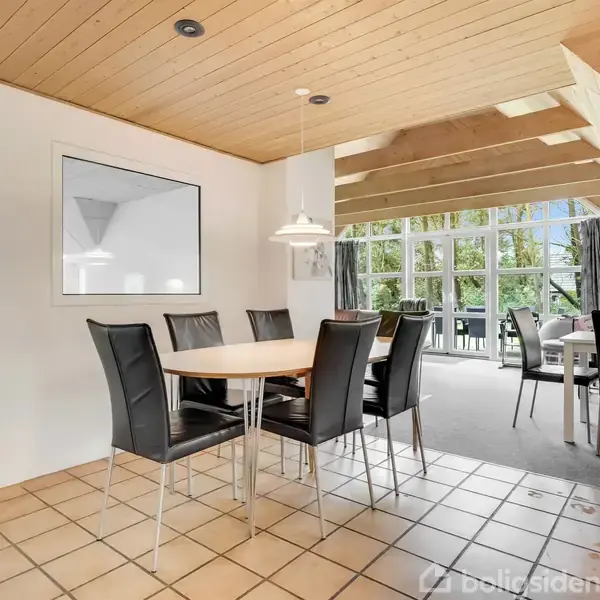 Spisebord med sorte stole står på flisegulv under træloft. Store vinduer i baggrunden giver udsigt til have, mens en lampe hænger over bordet i en lys og åben spisestue.