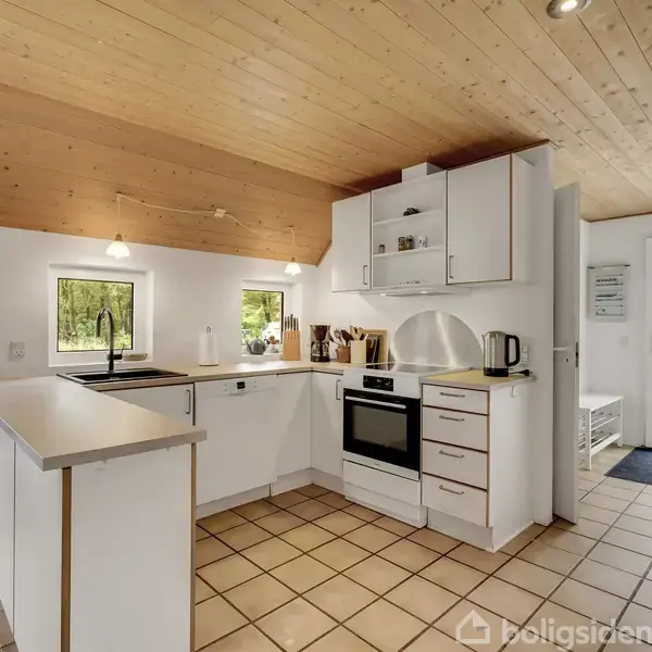 Et hvidt, moderne køkken står med komfur og vask, omgivet af hvide skabe. Trægulv og loft skaber en hyggelig atmosfære. Udenfor vinduet ses grønne træer.