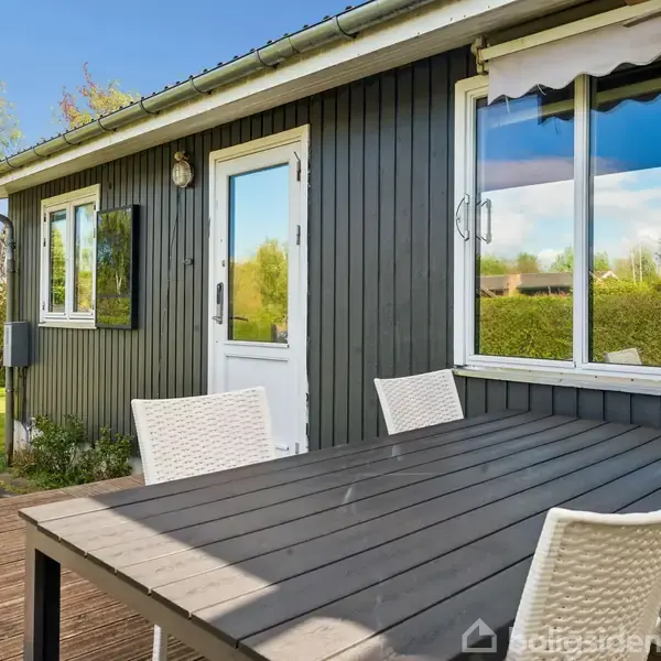 En sortmalet træbygning med store vinduer og en hvid dør. Foran står et bord med hvide stole. Omgivet af en grøn have under en blå himmel.