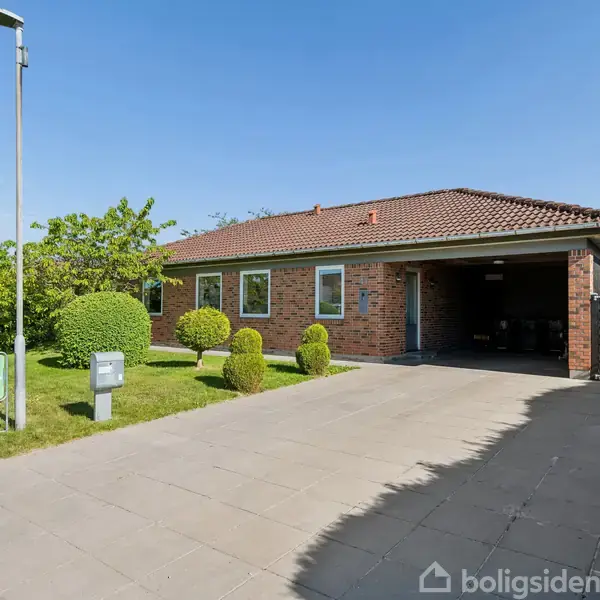 Murstenshus står på flisebelagt indkørsel med carport, omgivet af buske og et græsareal. En skilt ved vejen viser teksten "Ny bolig".